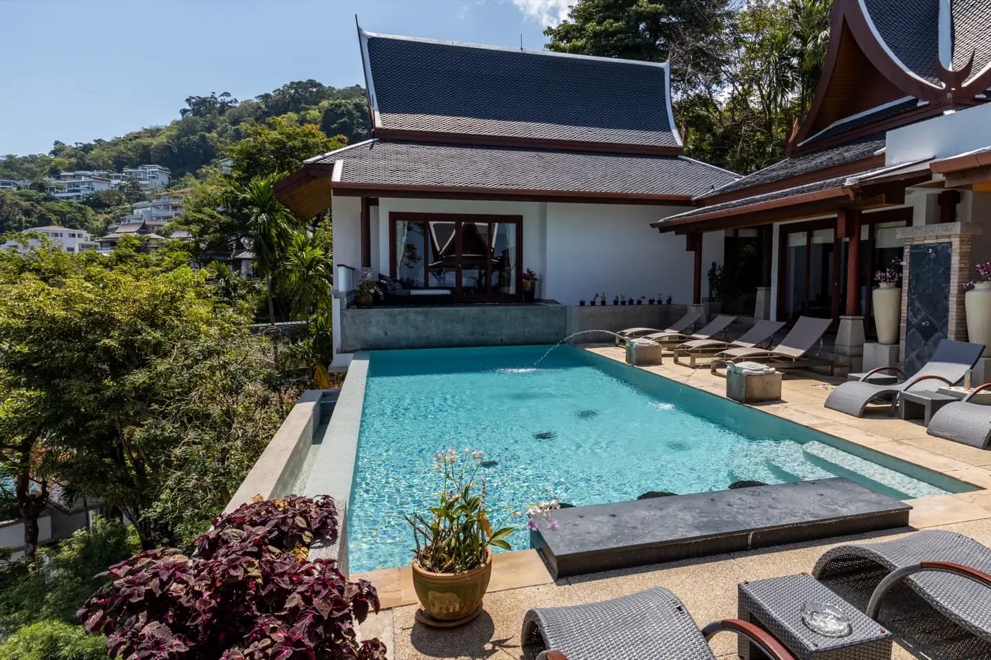Pool terrace with loungers and potted plants below a Thai-roofed house