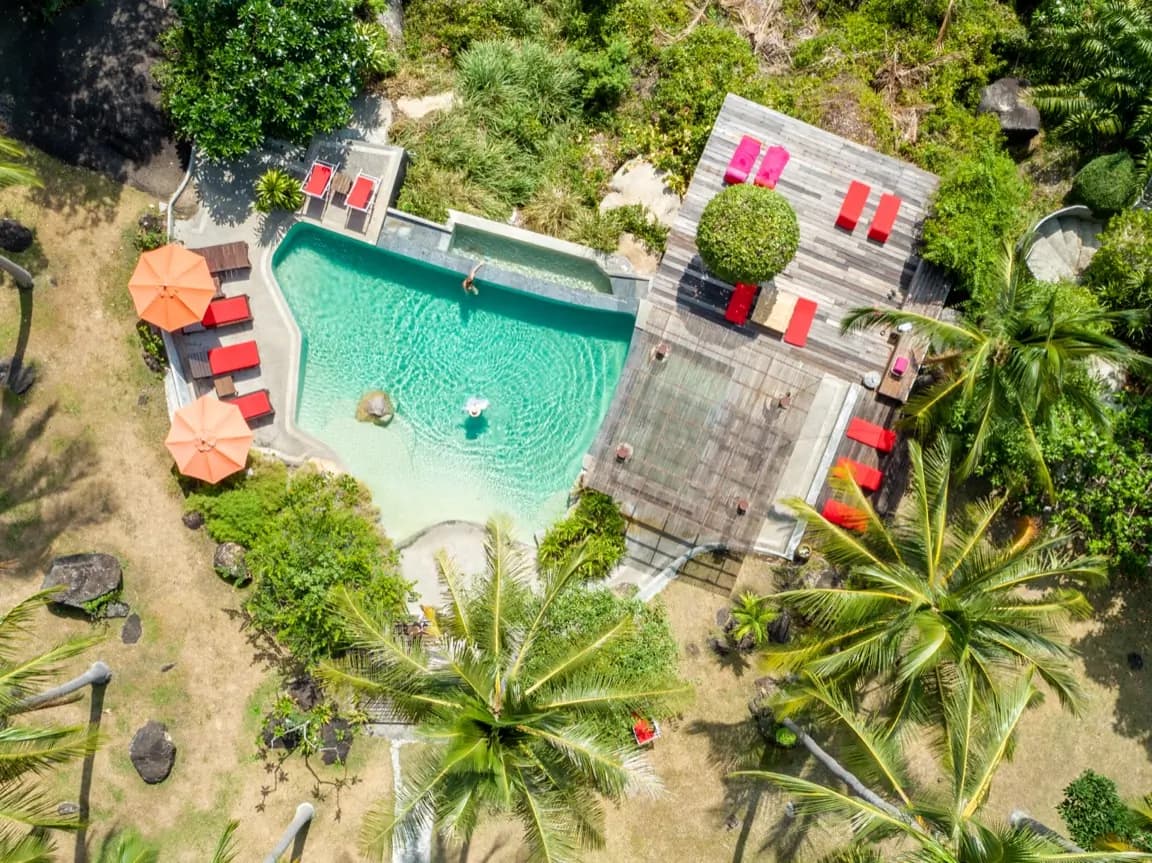 Top-down drone view of a lagoon pool among palms, sun decks and red loungers