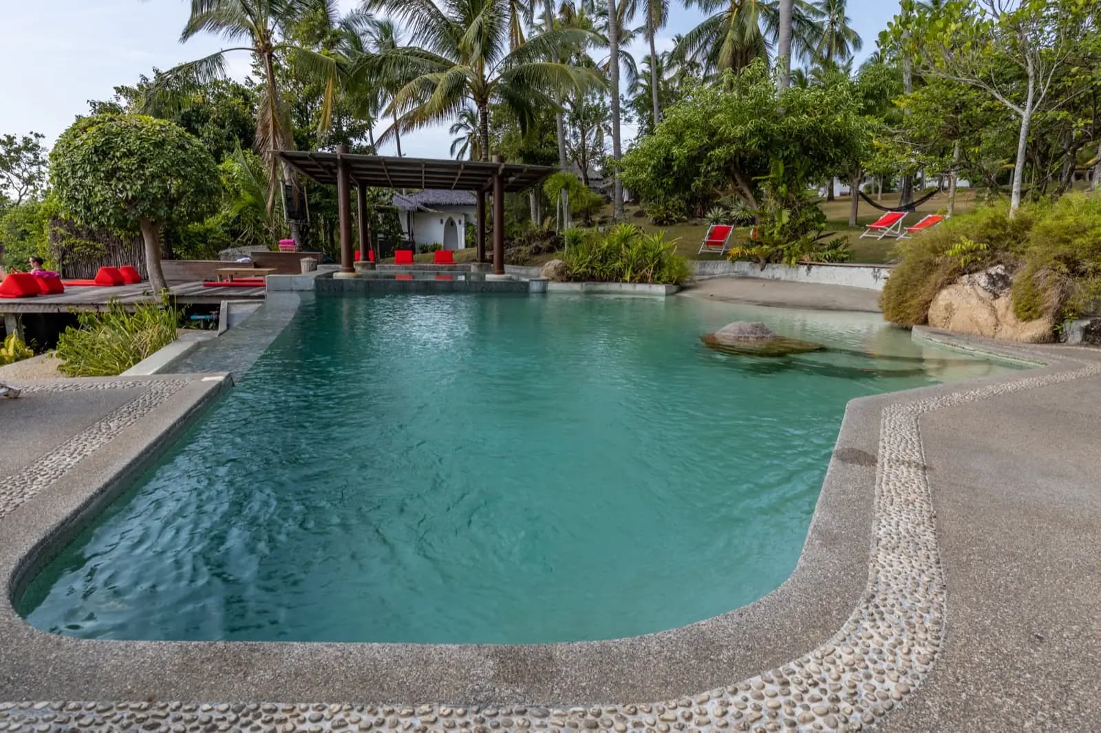 Curving resort pool with a pebble-mosaic edge and a pergola among tropical gardens