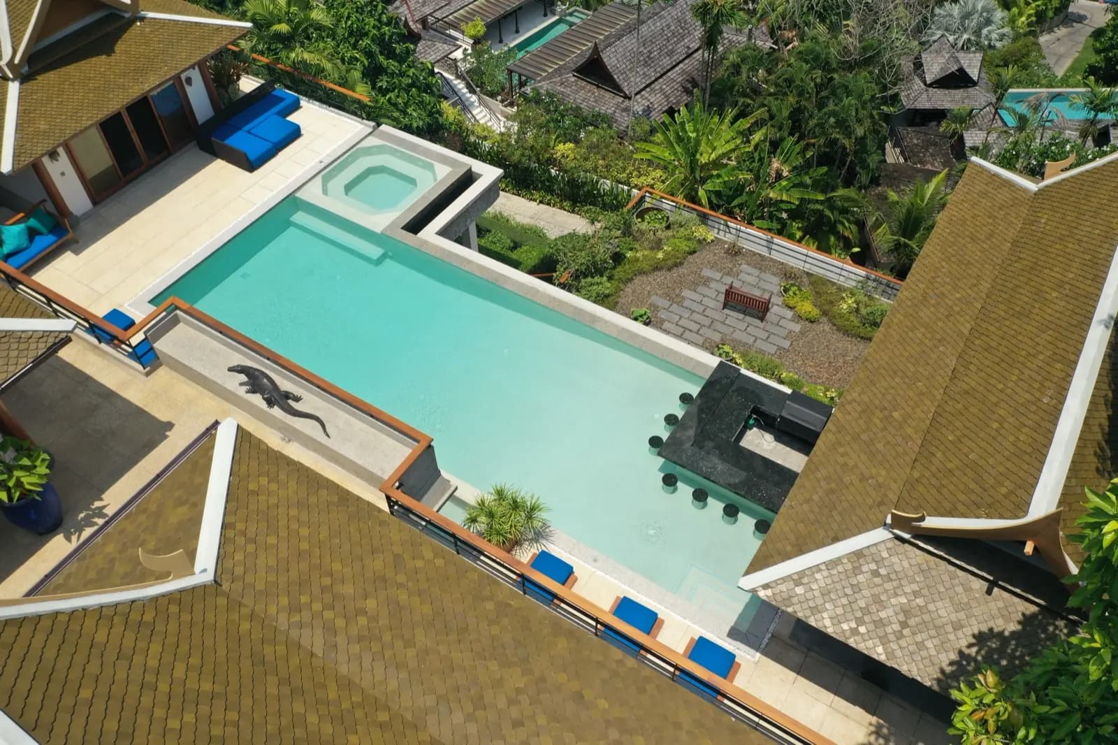 Diagonal overhead view of a turquoise pool between golden Thai rooftops and tropical garden