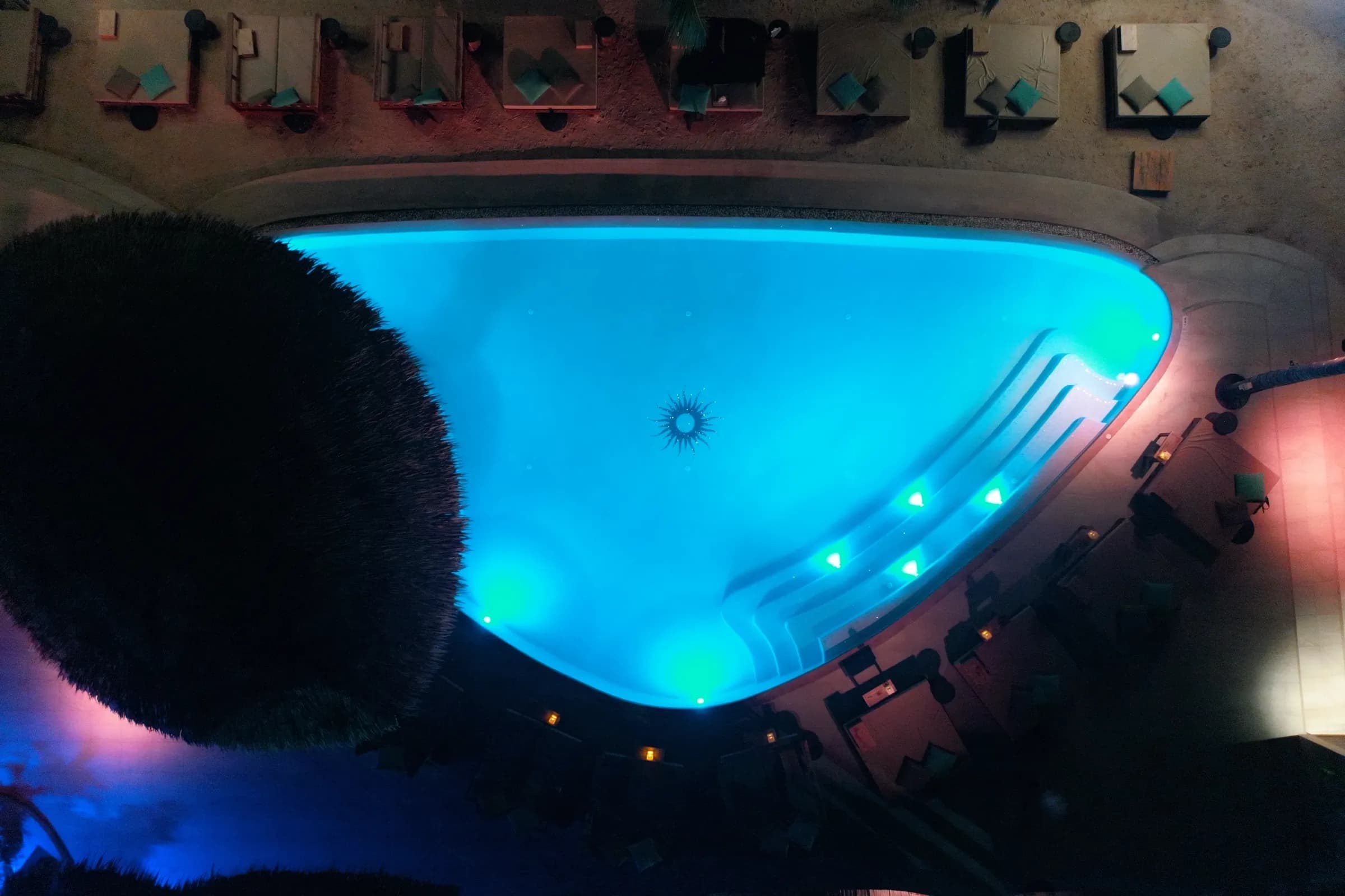 Overhead night view of the pool glowing electric cyan, with lit steps and the Carpe Diem logo set in the floor