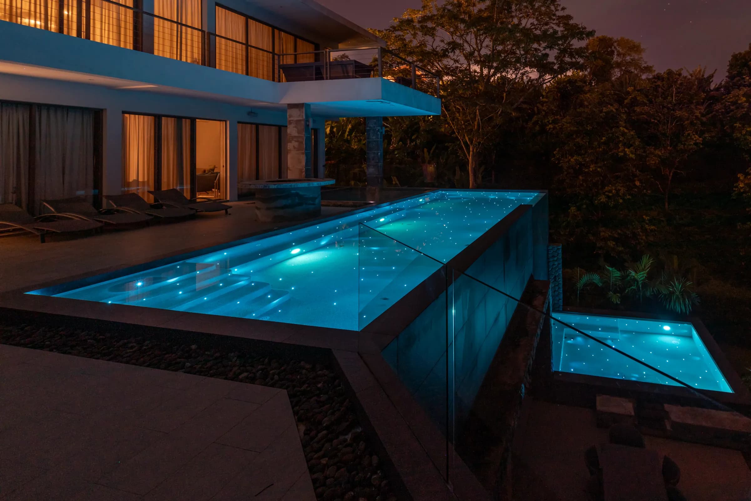 Raised overflow lap pool glowing cyan with fibre-optic starlight in front of a lit hillside villa at dusk