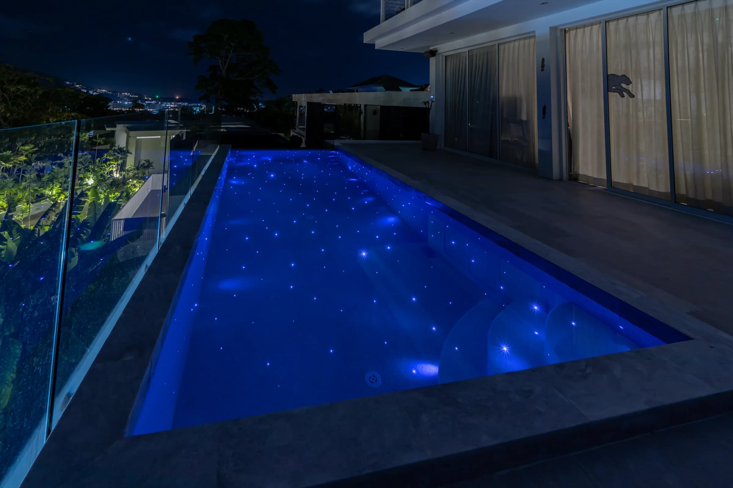 Glass-fenced terrace lap pool lit deep blue with starlight points and city lights beyond