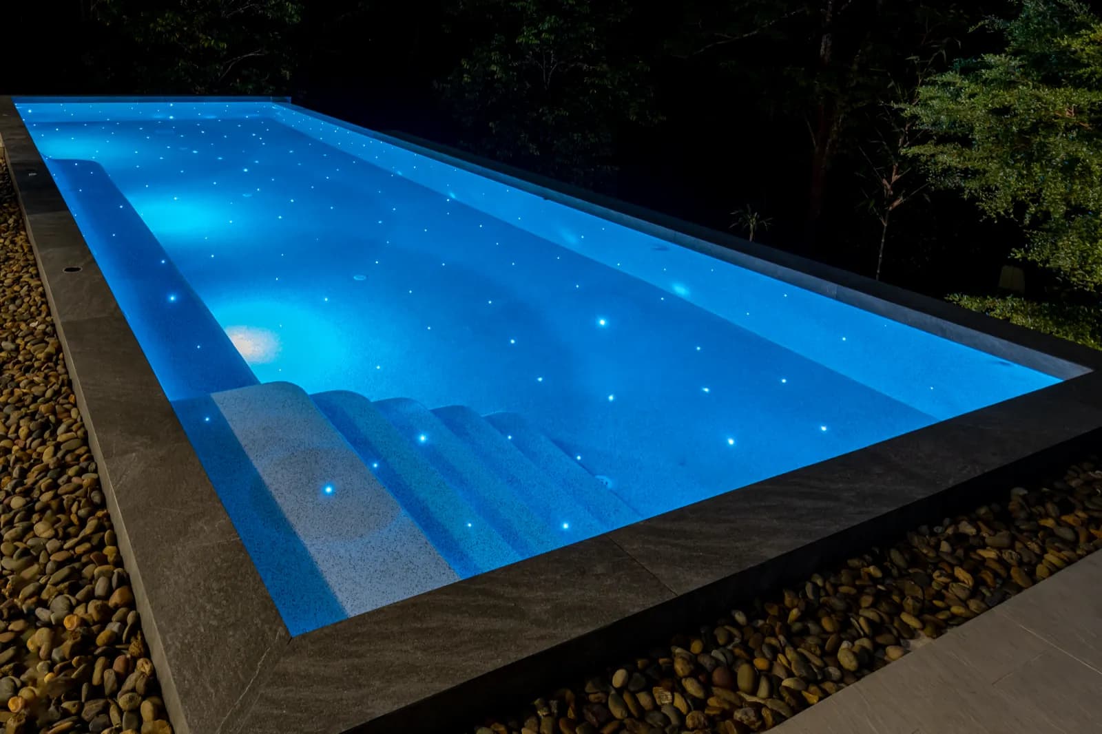 Close view of glowing pool steps and pebble finish ringed by a river-stone border at night