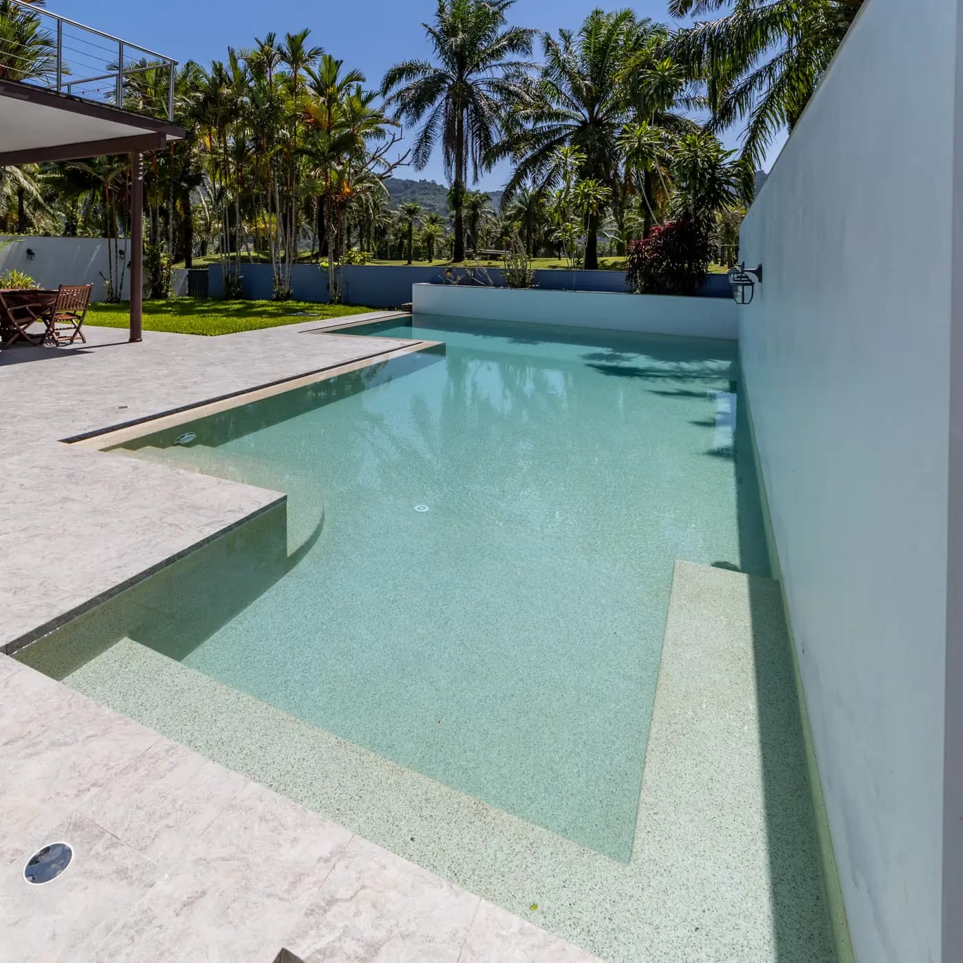 Pale pebble pool with a shallow sun-shelf beside a white feature wall and palms