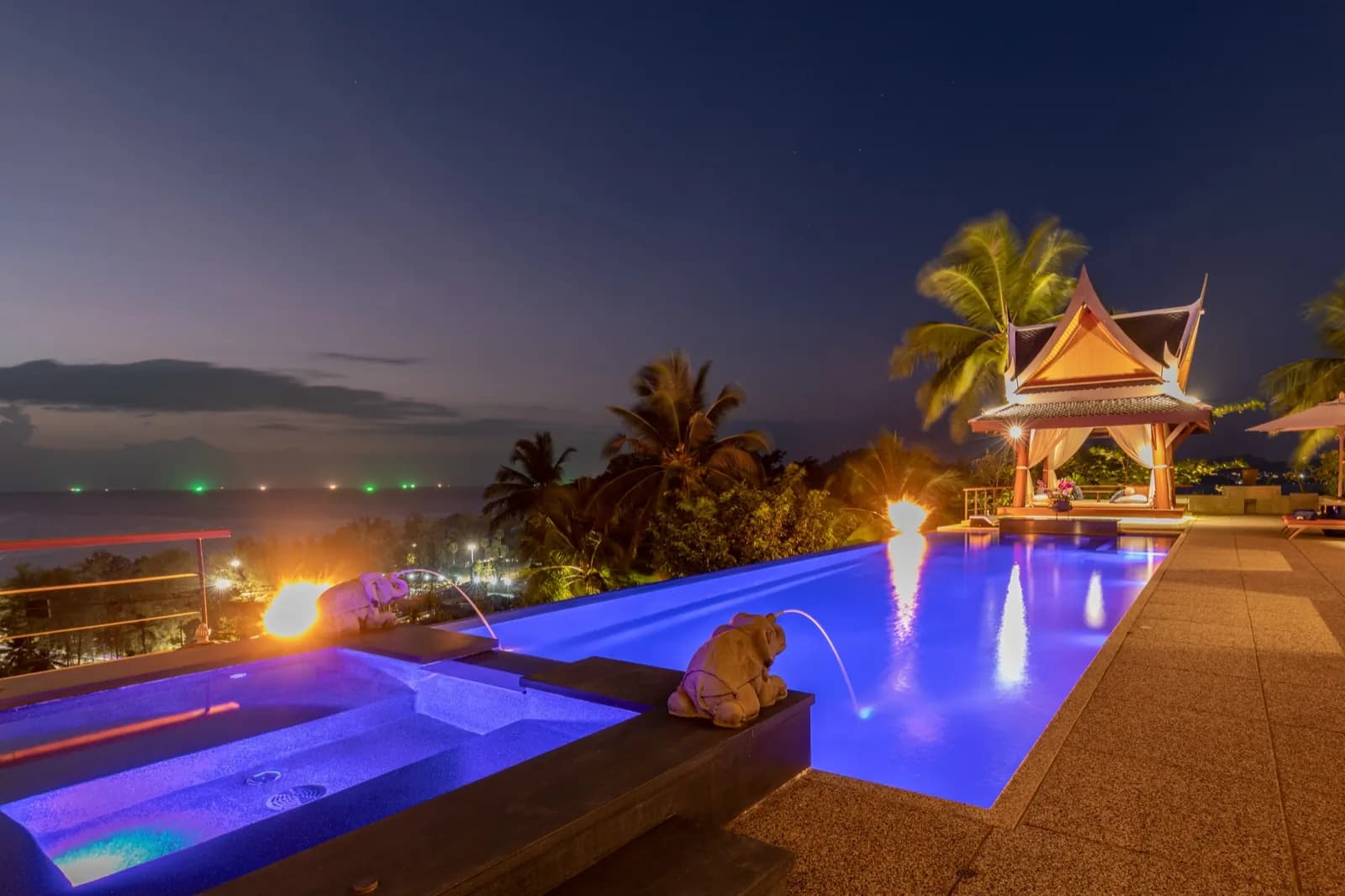 Violet-lit pool, fire features and Thai sala against the evening sea