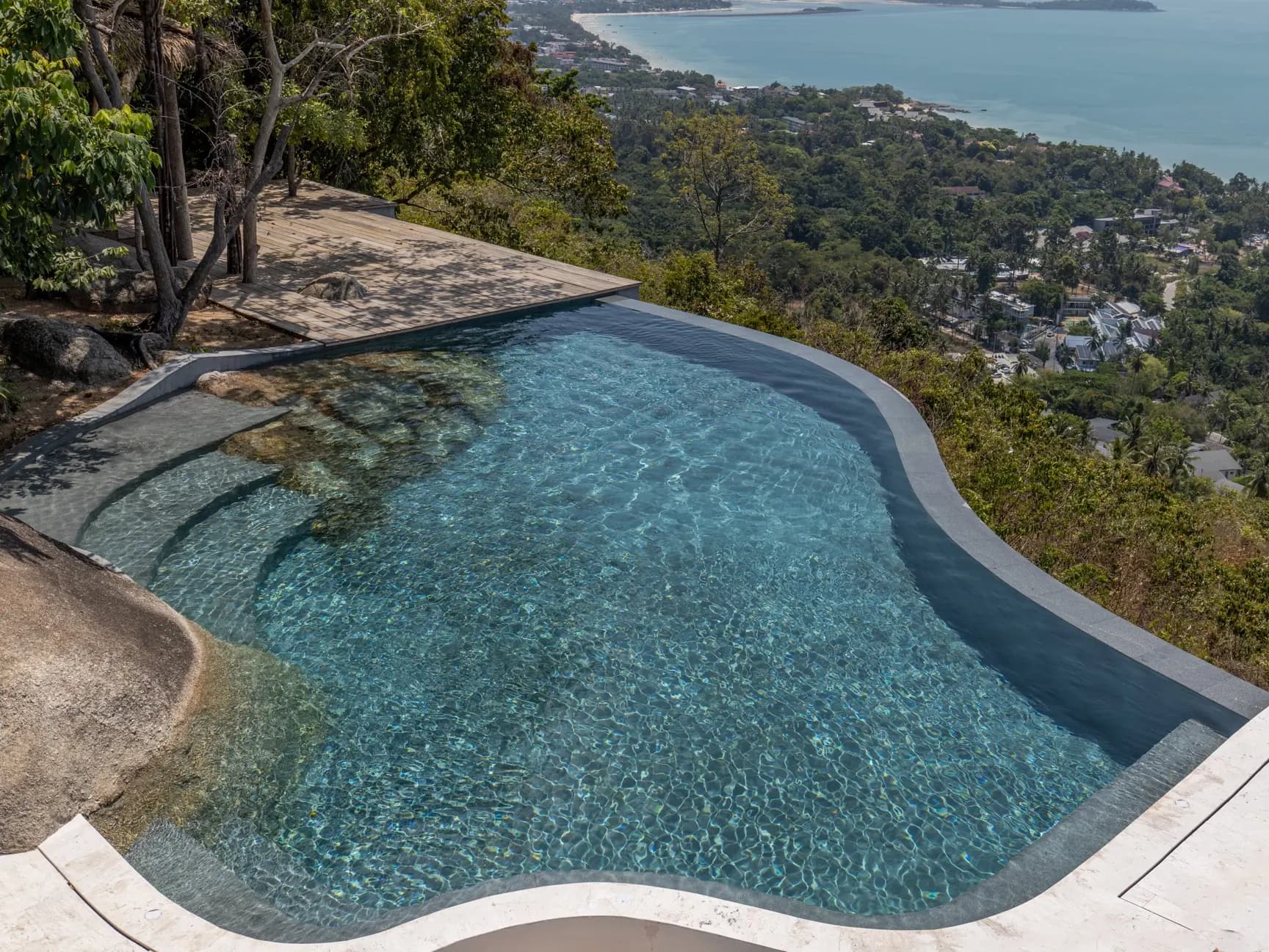 Curved infinity pool wrapping a granite boulder above a forested hillside and bay