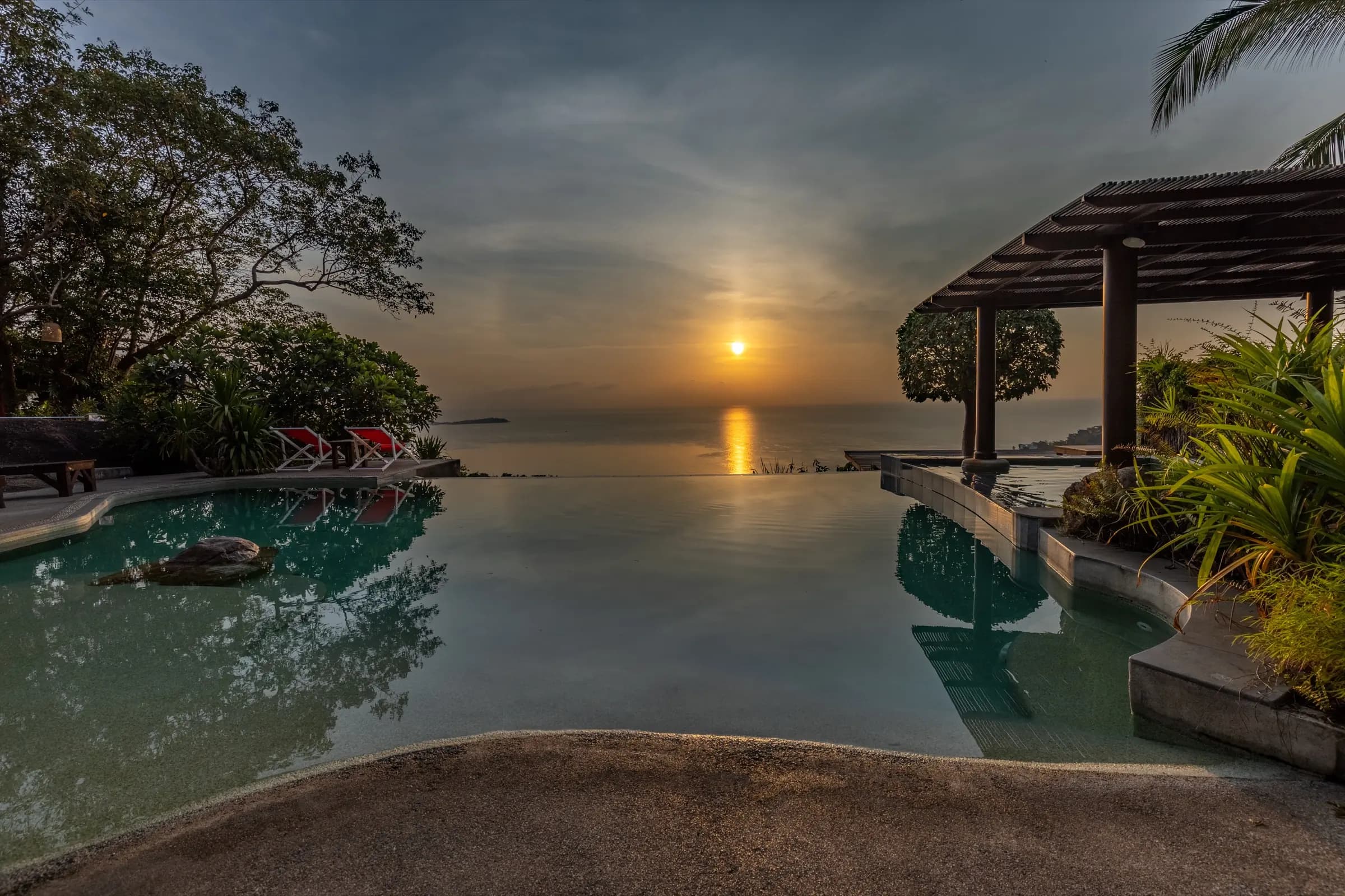 Free-form infinity pool reflecting the sunset over the Gulf of Thailand beside a timber pergola