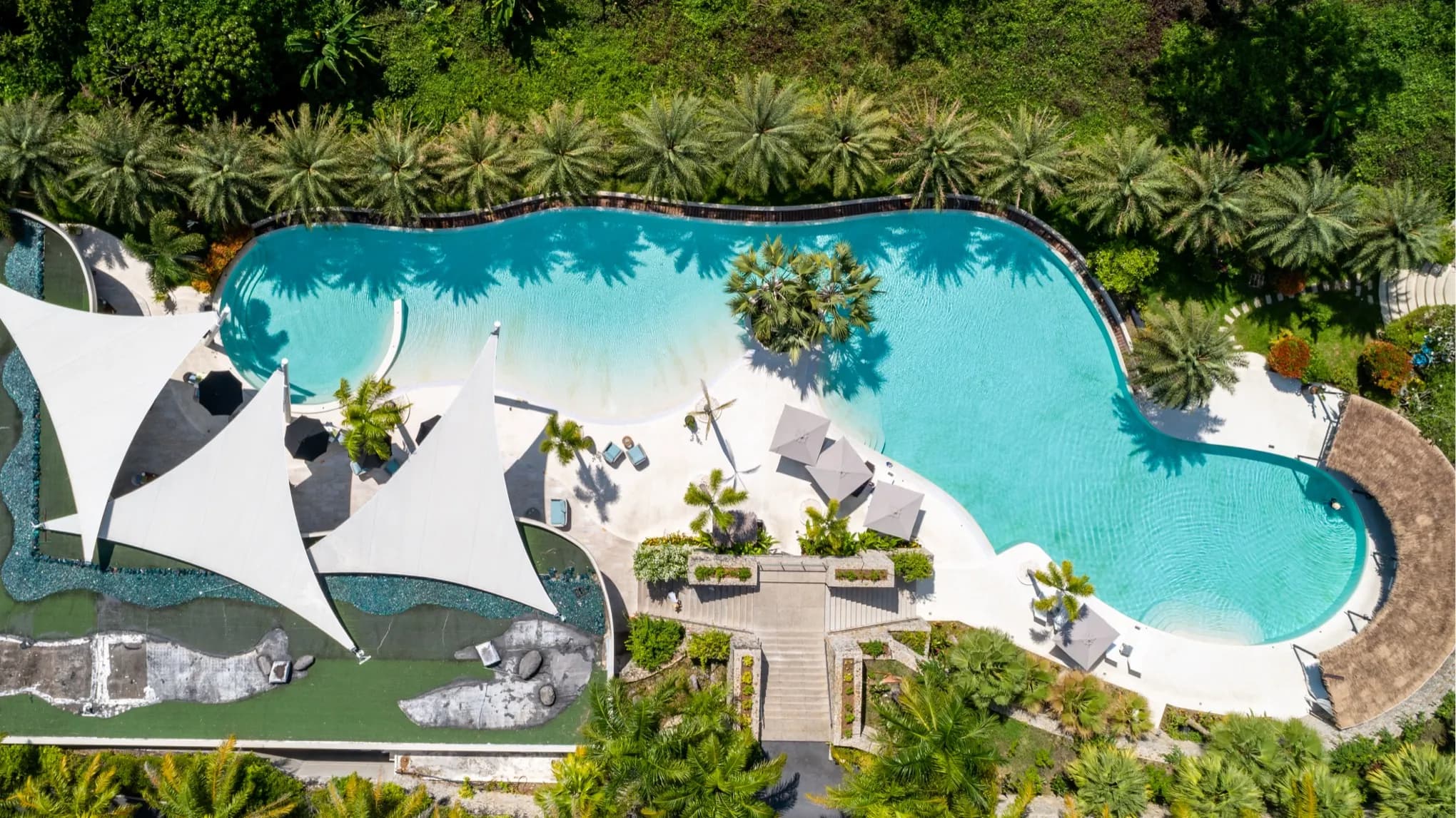 Aerial view of a free-form lagoon pool with white sand entry and sail-shade canopies